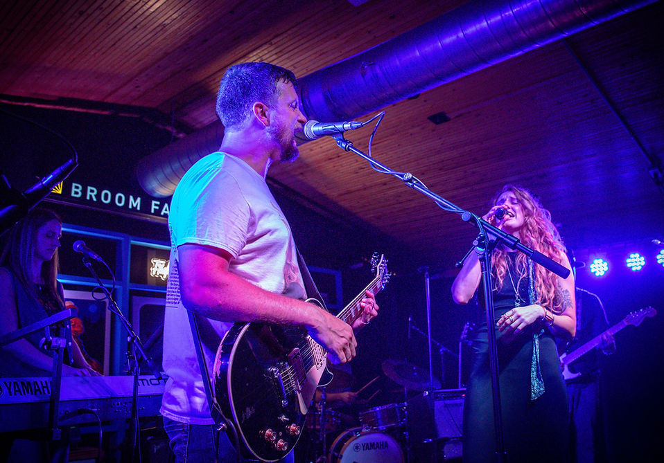 Evan Massey (left) strums a guitar and sings into microphone while looking at Krista Van Slingerland as she sings while clutching a microphone during a live performance at Broom Factory in Kingston.