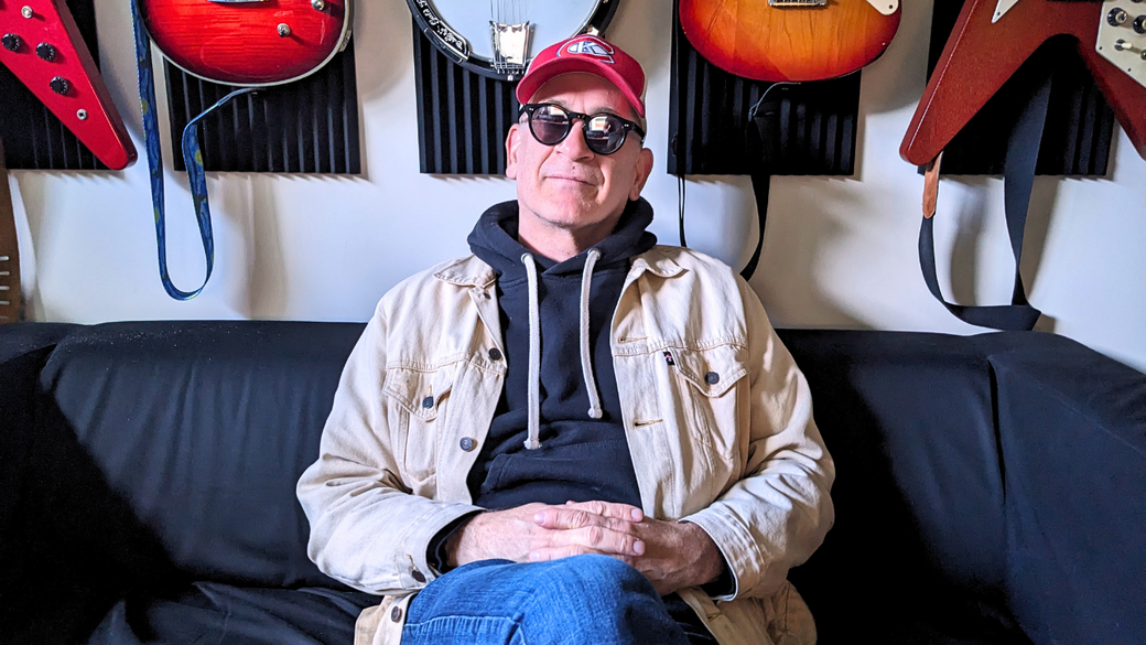 Steve Jordan sits on a black sofa in front of several guitars hanging on the wall, with his legs crossed and hands in his lap, wearing mirrored sunglasses and a red Kingston Canadians hockey team ball cap. 